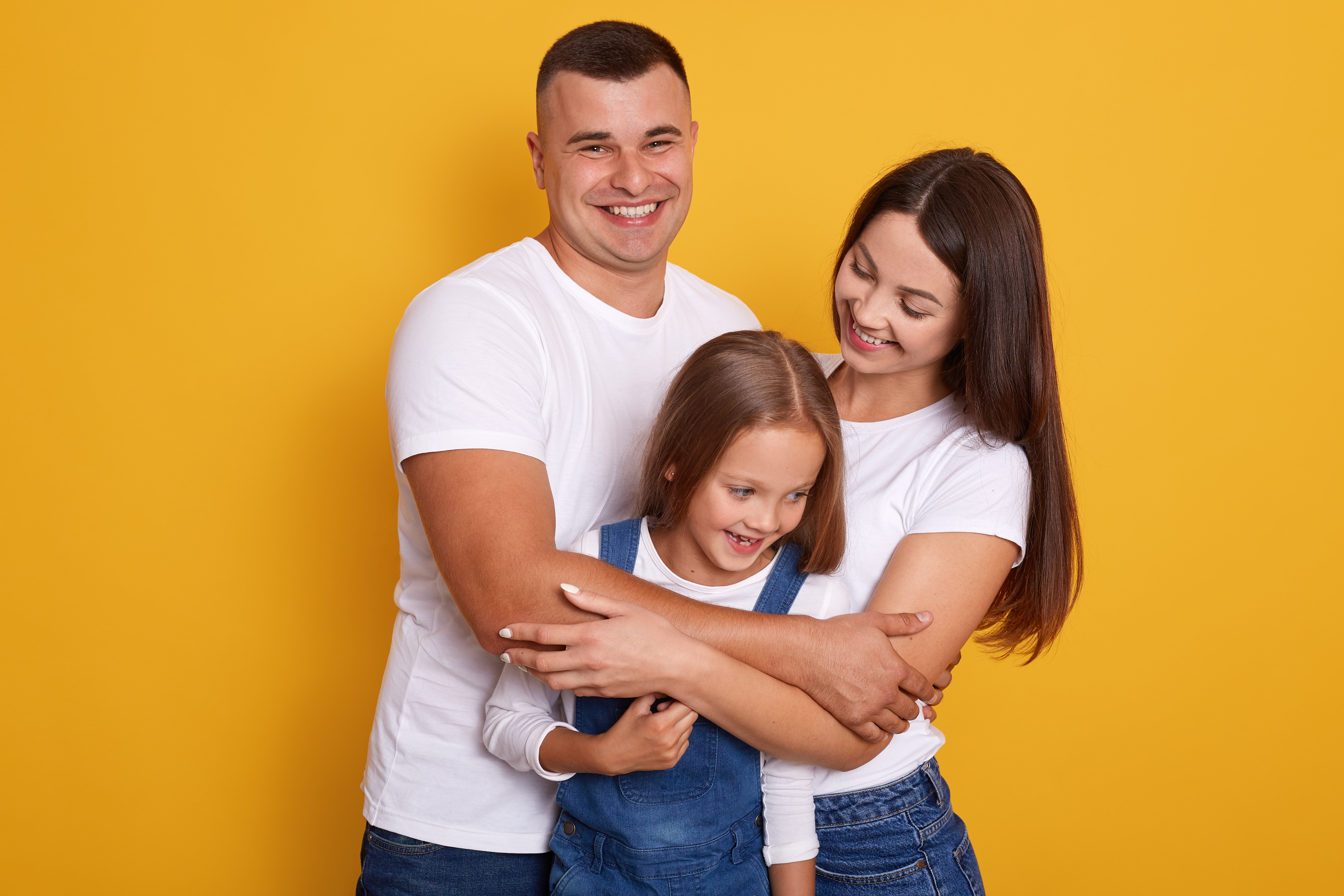 Mom, dad, and daughter all wearing white tshirts and jeans standing against a yellow background.